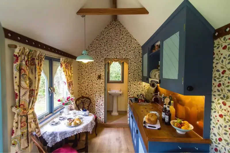 The kitchen and dining corner with blue cabinets, floral wallpaper and a lace-covered table