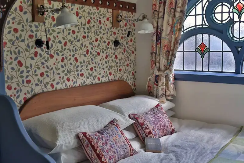 The double bed with embroidered cushions, floral wallpaper and stained glass